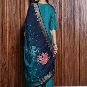 May include: A teal-colored, three-quarter sleeve outfit with matching pants. A dark blue shawl with a floral pattern and gold trim is draped over the shoulders. The outfit is worn with embellished flats.