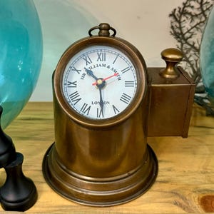 Op de afbeelding: Een vintage messing klok met een witte wijzerplaat en zwarte Romeinse cijfers. De wijzerplaat leest "Sir William & Smith London". De klok heeft een klein handvat aan de bovenkant en een rechthoekig zijstuk. De klok staat op een ronde basis.