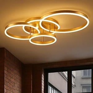 Könnte beinhalten: Eine moderne Deckenleuchte mit goldener Oberfläche, die aus mehreren miteinander verbundenen kreisförmigen LED-Leuchten besteht. Die Leuchte ist an einer hellen Decke montiert, mit einer Backsteinmauer und einem Fenster im Hintergrund.