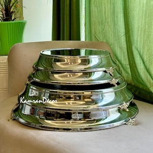 May include: A stack of four tiered, silver-colored serving platters. Each platter has a scalloped edge and decorative floral accents. The platters are arranged on a neutral-colored surface, with a green curtain in the background.