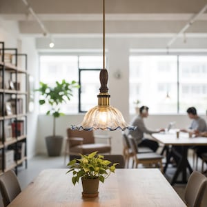 May include: A pendant light fixture featuring a clear, fluted glass shade with a ruffled edge and a subtle blue tint. The shade is connected to a dark wood neck and a brass-toned metal fitting, suspended by a thin, straight cord from the ceiling.