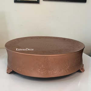May include: A copper-colored cake stand with a round top and ornate floral detailing. The stand has a textured surface and is supported by decorative feet. The text "KamranDecor" is visible on the side.