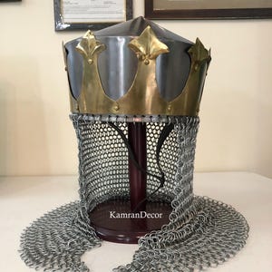 Puede incluir: Un casco de estilo medieval con una corona de metal plateado y una corona de color latón. El casco presenta un protector de cuello de cota de malla y se exhibe sobre un soporte de madera. El texto "KamranDecor" es visible.