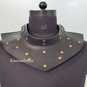 Può includere: Colletto in pelle nera con borchie color oro, esposto su un manichino nero. Il colletto ha un collo rotondo e un davanti svasato e appuntito. Il numero "42" è stampato sul manichino. Il testo "KamranDecor" è visibile.