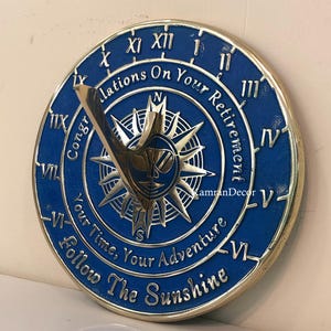 Könnte beinhalten: Ein blau-goldenes Ruhestands-Sonnenuhr mit römischen Ziffern und dem Text "Glückwünsche zu Ihrem Ruhestand, Ihre Zeit, Ihr Abenteuer, Folgen Sie dem Sonnenschein". Die Sonnenuhr hat ein Sonnen- und Kompassdesign.