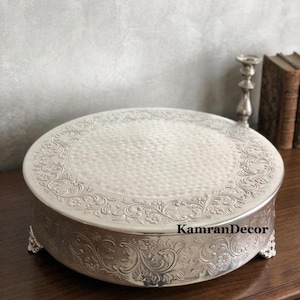 May include: Silver-colored, round cake stand with ornate floral and scrollwork detailing. The stand has a textured surface and is supported by small feet. The brand name "KamranDecor" is visible on the side.