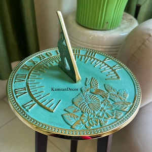 Könnte beinhalten: Ein teal-farbenes und goldenes Sonnenuhr mit römischen Ziffern und floralen Details. Die Sonnenuhr hat einen zentralen Gnomon und ist aus Metall gefertigt. Die Sonnenuhr ist ein dekoratives Gartenelement und trägt den Text "KamranDecor".