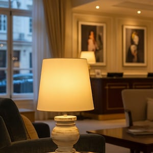 May include: A lit table lamp with a cream-colored shade and a distressed white base. The lamp sits on a dark brown table in a room with a window and artwork on the wall. The lamp's warm glow illuminates the surrounding area.
