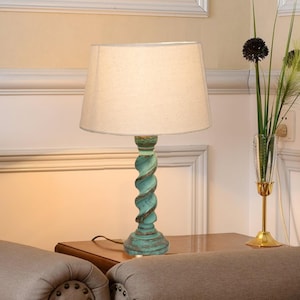 Puede incluir: Una lámpara de mesa decorativa con una pantalla beige y una base torcida turquesa. La lámpara está sobre una mesa de madera. La base tiene un acabado envejecido, lo que le da un atractivo vintage. Un elemento decorativo para el hogar.