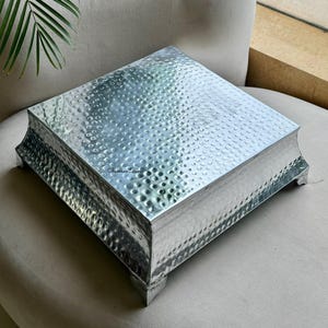 Könnte beinhalten: Ein quadratischer, silberfarbener Kuchenständer aus Metall mit gehammerter Textur. Der Ständer hat eine leicht erhöhte Basis mit dekorativen Füßen. Die obere Oberfläche ist flach und die Kanten sind leicht gebogen.