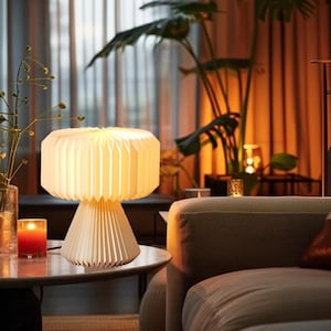 May include: A cream-colored, pleated paper table lamp with a conical base and a cylindrical shade. The lamp is on a round table with a lit red candle. The background includes a sofa, plants, and a window.