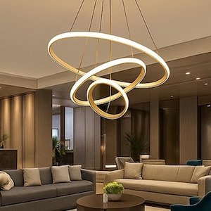 Puede incluir: Lámpara colgante moderna dorada con tres anillos circulares iluminados e interconectados. La lámpara cuelga del techo con finos cables, emitiendo un brillo cálido. Adecuada para interiores contemporáneos.