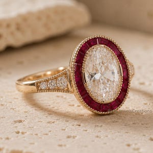 Op de afbeelding: Een gouden ring met een grote ovaal geslepen middensteen omringd door een halo van dieprode robijnen in baguette-slijpvorm. De band is gemaakt van gepolijst goud en bevat drie kleine ronde diamanten aan weerszijden van de zetting.