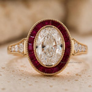 Puede incluir: Un anillo de oro con una gran piedra central blanca de corte ovalado rodeada por un halo de rubíes rojos de corte baguette. La banda incluye pequeñas piedras blancas redondas a cada lado con detalles milgrain. El texto BelleJewelsCreations es visible.