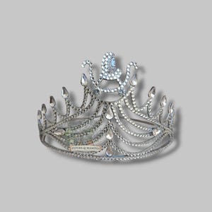 May include: A silver-toned tiara with clear crystal accents. The crown features a central design and multiple teardrop-shaped crystals. The base of the tiara has the text "PIPERS & MASONS".