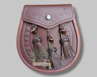 May include: A brown leather sporran with a decorative flap and three tassels. The sporran features embossed floral patterns and a silver-colored stud closure. The bottom of the sporran has a small image with the text "PIPERS & MASONS".