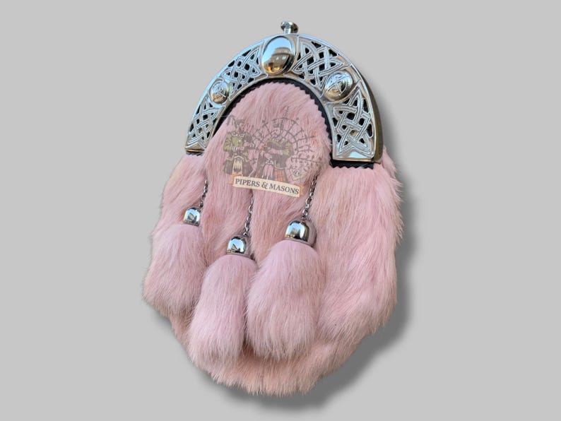 May include: A pink fur sporran with a silver Celtic design cantle. The sporran has three matching fur tassels with silver metal accents. The text "PIPERS & MASONS" is visible on the front.