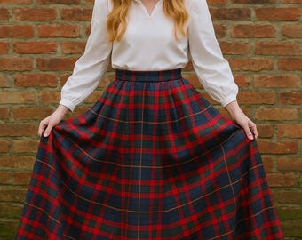 May include: A long, full plaid skirt in red, navy, and green, paired with a white blouse. The skirt features a classic tartan pattern. The model is standing in front of a brick wall.