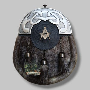 Puede incluir: Un sporran escocés tradicional con cuerpo de piel, detalles metálicos plateados y frente de cuero negro. Presenta un símbolo masónico y el texto "PIPERS & MASONS". Cadenas, borlas y una parte superior de metal decorativa.