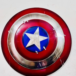 May include: A round shield with a red, silver, and blue color scheme. The shield features a red outer ring, a silver middle ring, and a blue center with a white star. The shield has a distressed appearance.