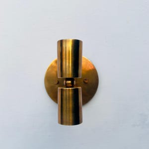 Op de afbeelding: Een vintage wandlamp met een messing afwerking. De armatuur heeft een ronde achterplaat en twee cilindrische lichthouders. De lamp is ontworpen om aan de muur te worden bevestigd en zorgt voor een warm, sfeervol licht.