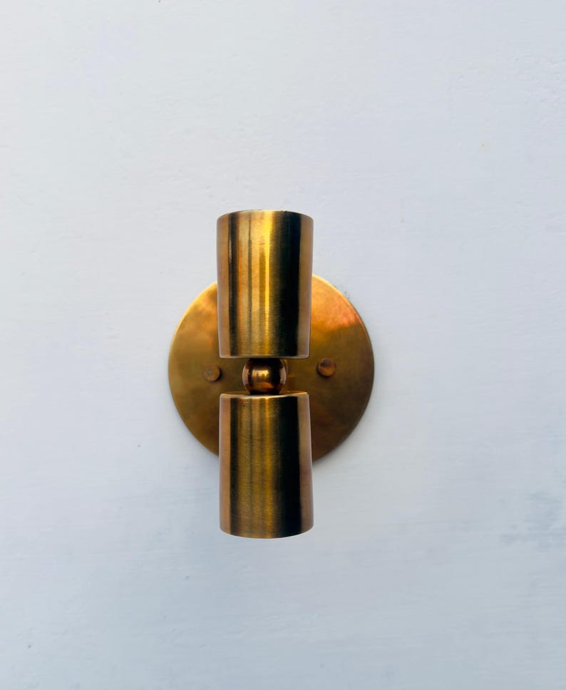 Op de afbeelding: Een vintage wandlamp met een messing afwerking. De armatuur heeft een ronde achterplaat en twee cilindrische lichthouders. De lamp is ontworpen om aan de muur te worden bevestigd en zorgt voor een warm, sfeervol licht.