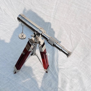 Puede incluir: Un telescopio plateado pulido con un trípode rojo y plateado. El telescopio tiene un diseño detallado y está unido a una pequeña cadena plateada con una pieza circular. Las patas del trípode son de color rojo intenso.