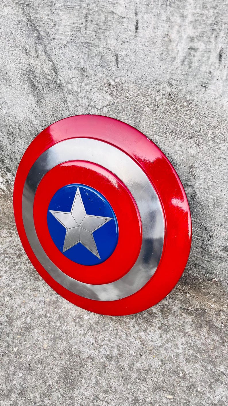 May include: A red, white, and blue shield with a silver star in the center. The shield has a circular design with a red outer ring, a silver middle ring, and a blue center. A classic superhero accessory.
