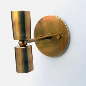 Op de afbeelding: Een messing wandlamp met een cilindrische lamp en een ronde achterplaat. De lamp heeft een warme, metallic afwerking en is op een witte muur gemonteerd. De lamp bestaat uit twee gestapelde cilinders.