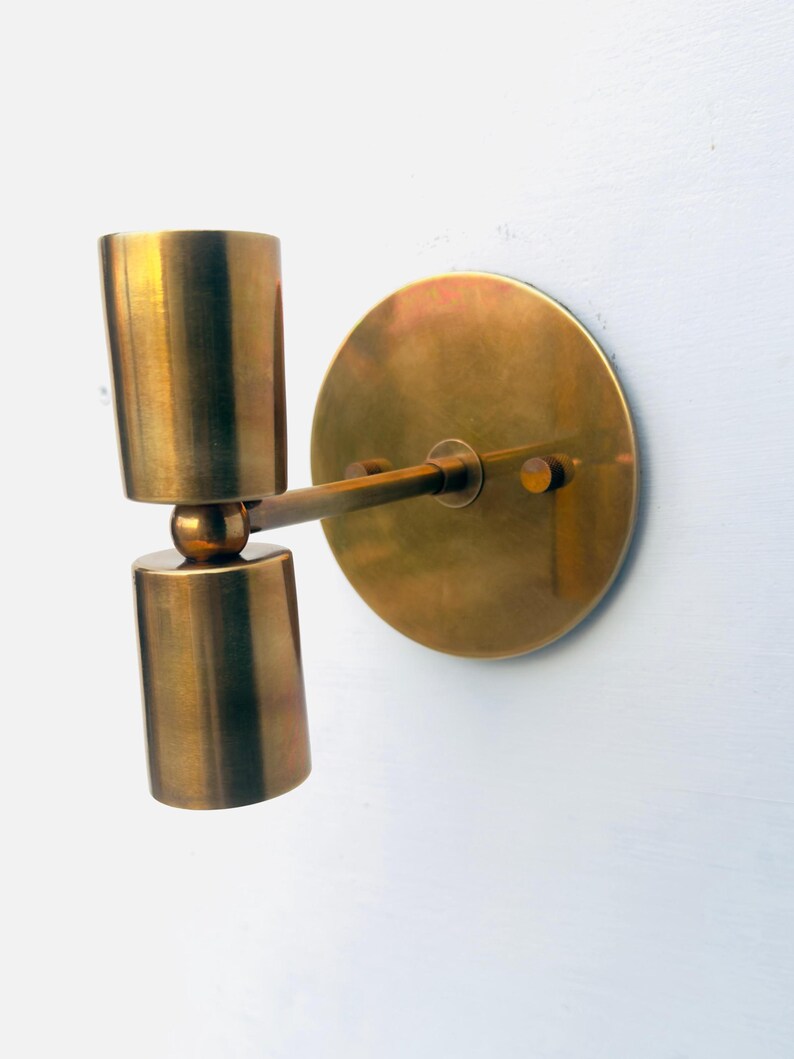 Op de afbeelding: Een messing wandlamp met een cilindrische lamp en een ronde achterplaat. De lamp heeft een warme, metallic afwerking en is op een witte muur gemonteerd. De lamp bestaat uit twee gestapelde cilinders.
