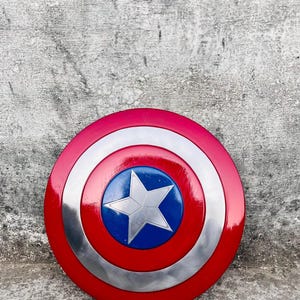 May include: A round shield with a red, white, and blue color scheme. The shield has a red outer ring, a silver middle ring, and a blue center with a white star. The shield is a replica of Captain America's shield.