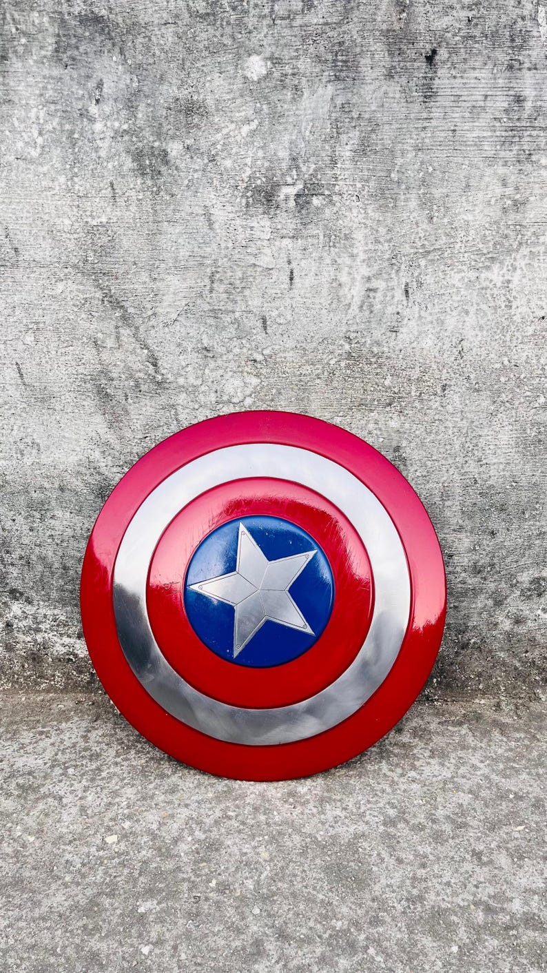 May include: A round shield with a red, white, and blue color scheme. The shield has a red outer ring, a silver middle ring, and a blue center with a white star. The shield is a replica of Captain America's shield.