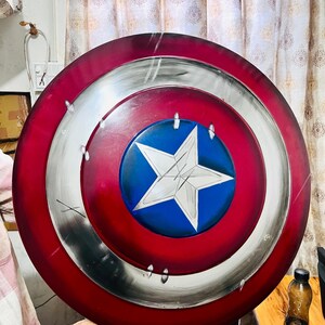 May include: A round shield with a red, white, and blue color scheme. The shield features a central blue circle with a white star, surrounded by concentric rings of red and silver. The shield is a replica of Captain America's shield.