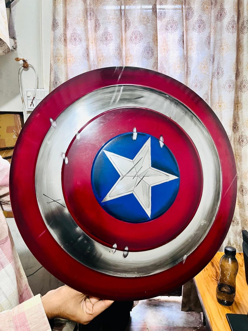 May include: A round shield with a red, white, and blue color scheme. The shield features a central blue circle with a white star, surrounded by concentric rings of red and silver. The shield is a replica of Captain America's shield.
