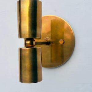 Op de afbeelding: Een messing wandlamp met een cilindrisch ontwerp. De lamp heeft twee gestapelde cilinders die zijn verbonden door een kogelgewricht, gemonteerd op een cirkelvormige achterplaat. De armatuur heeft een warme, metallic afwerking.
