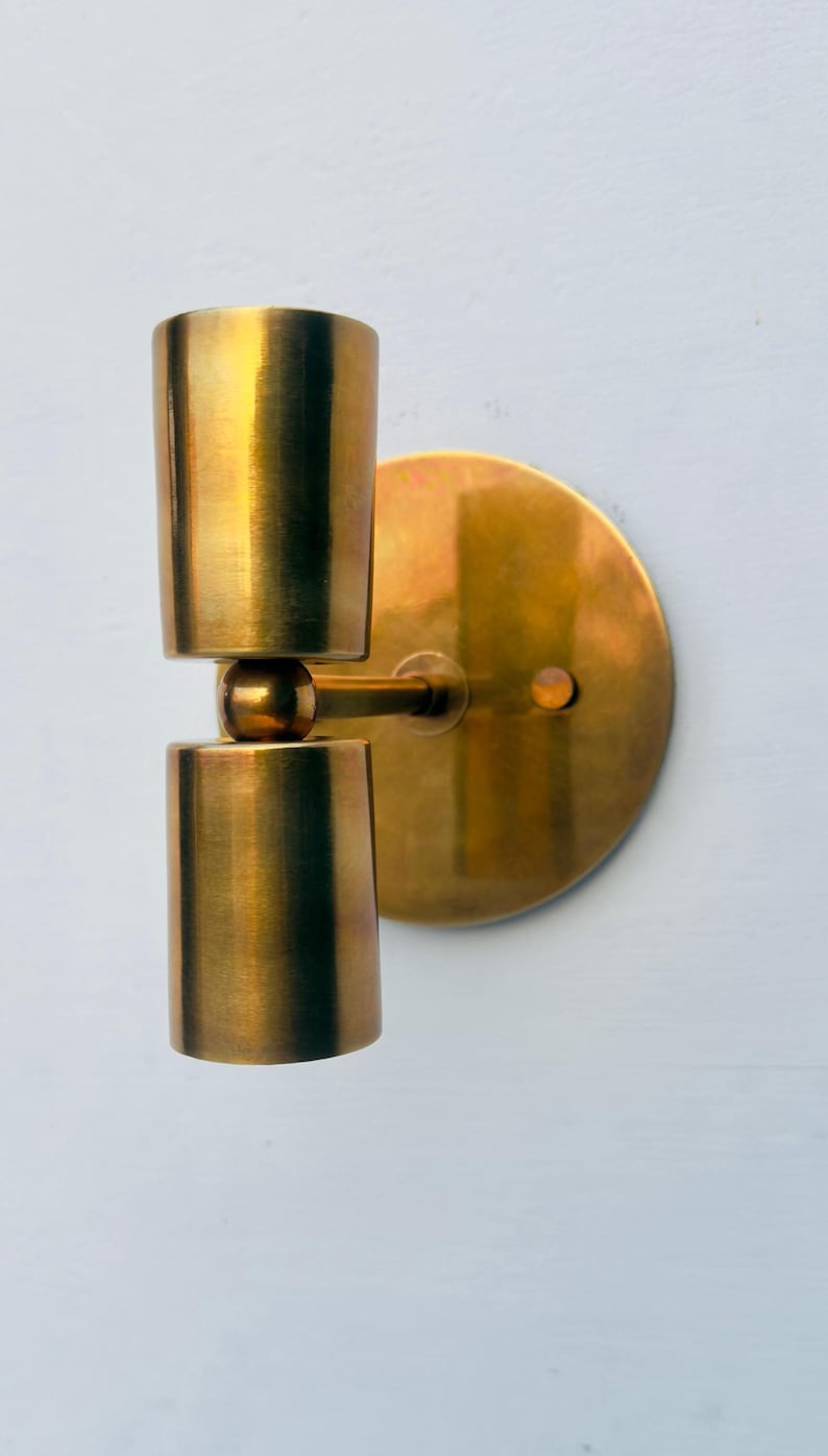Op de afbeelding: Een messing wandlamp met een cilindrisch ontwerp. De lamp heeft twee gestapelde cilinders die zijn verbonden door een kogelgewricht, gemonteerd op een cirkelvormige achterplaat. De armatuur heeft een warme, metallic afwerking.