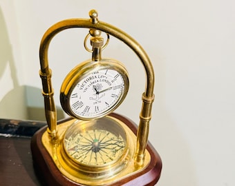 May include: A brass and wood desk clock with a compass and a pocket watch. The pocket watch is white with black Roman numerals and the words "Victoria London 1876". The compass is gold with a glass cover. The base is dark brown.
