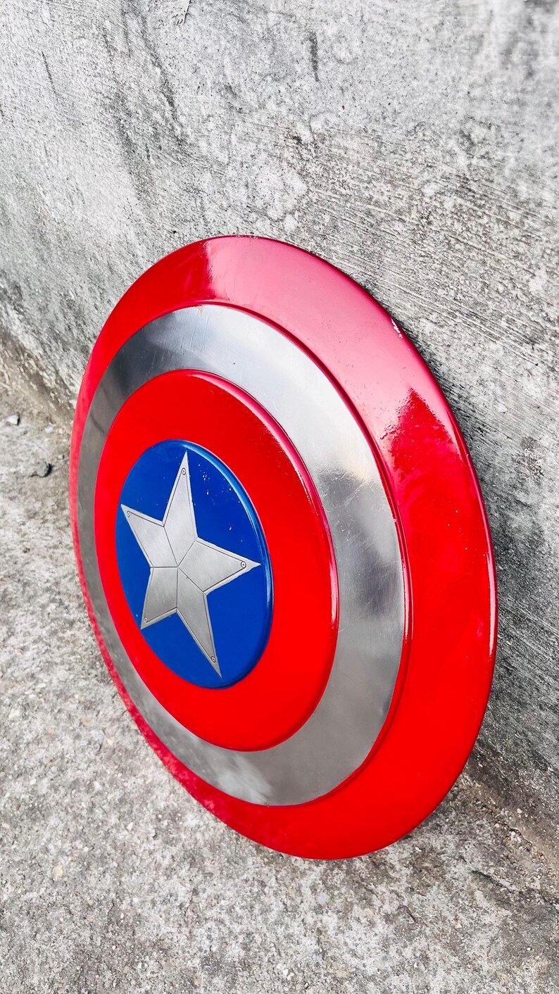 May include: A red, white, and blue Captain America shield. The shield has a red outer ring, a silver middle ring, and a blue center with a white star. The shield is round and appears to be made of metal.