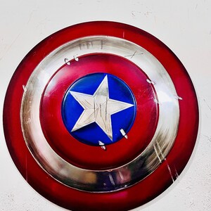 May include: A round shield with a red, silver, and blue color scheme. The shield features a large white star on a blue circle in the center. The outer ring is red, and the middle ring is silver. The shield has a distressed look.
