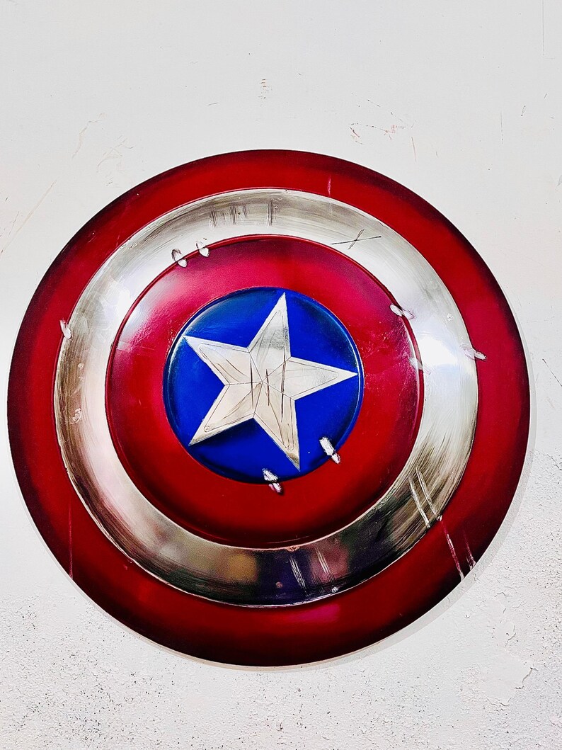 May include: A round shield with a red, silver, and blue color scheme. The shield features a large white star on a blue circle in the center. The outer ring is red, and the middle ring is silver. The shield has a distressed look.