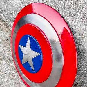 May include: A red, white, and blue Captain America shield. The shield has a red outer ring, a silver middle ring, and a blue center with a white star. The shield is round and appears to be made of metal.