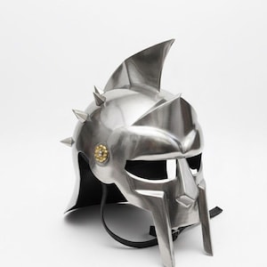 Könnte beinhalten: Silberfarbener Gladiatorenhelm mit Flosse und Stacheln. Der Helm hat einen Gesichtsschutz und ein goldfarbenes Emblem. Ein schwarzer Riemen ist befestigt. Metallhelm.