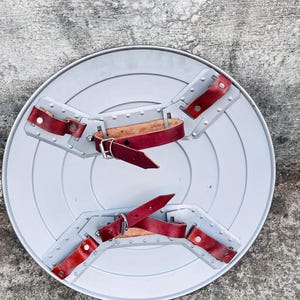 May include: A gray, circular shield with a series of concentric rings. It features red leather straps and silver rivets for securing the shield. The shield is designed with a handle and arm straps for easy carrying.