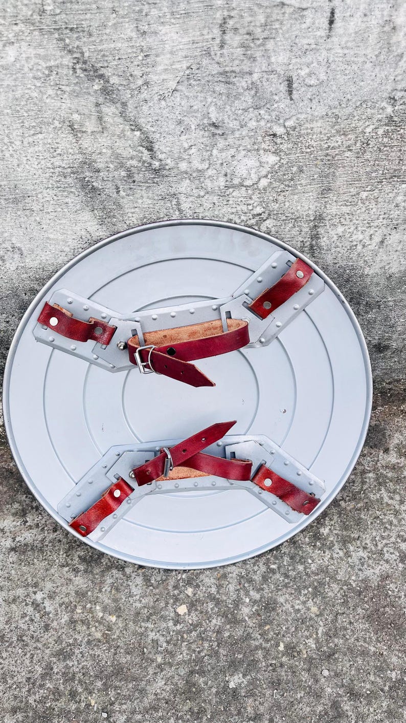 May include: A gray, circular shield with a series of concentric rings. It features red leather straps and silver rivets for securing the shield. The shield is designed with a handle and arm straps for easy carrying.