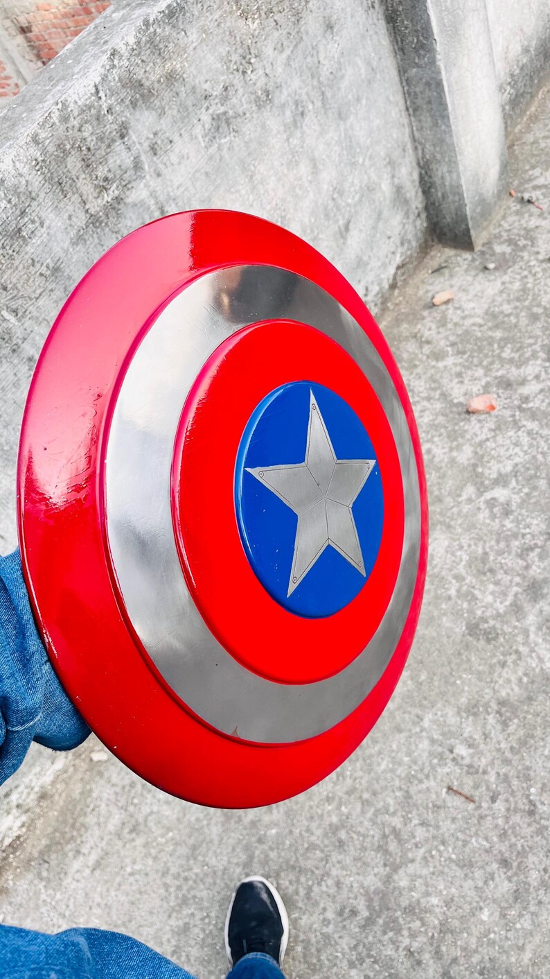 May include: A red, silver, and blue Captain America shield. The shield has a red outer ring, a silver middle ring, and a blue center with a silver star. The shield is round and appears to be made of metal.