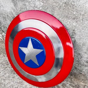 May include: A red, silver, and blue shield with a star emblem. The shield has a circular design with a red outer ring, a silver middle ring, and a blue center with a silver star. This item is a collectible.