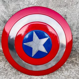 May include: A red, white, and blue shield with a silver star in the center. The shield has a circular design with concentric rings of red, silver, and blue. The shield is a prop or collectible.