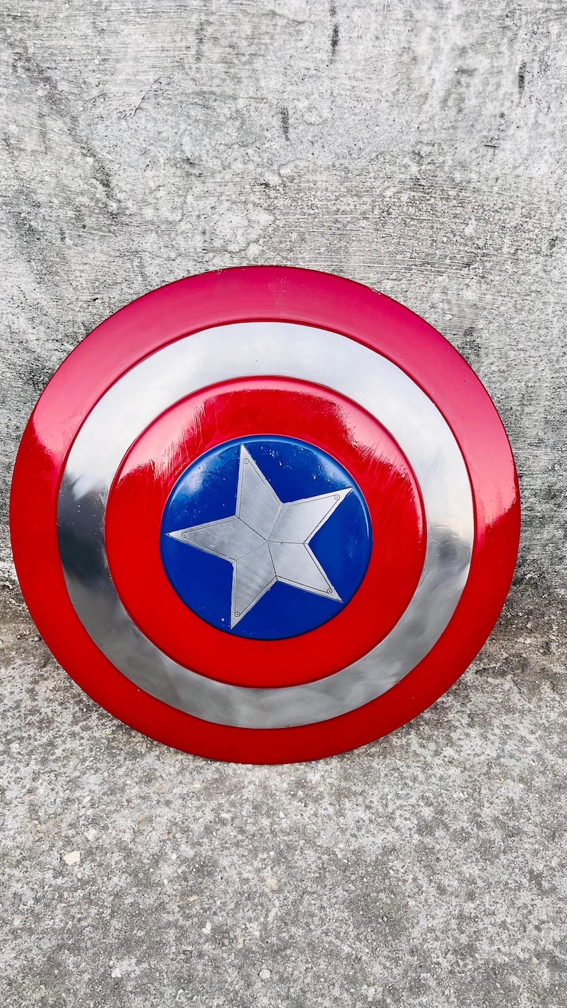 May include: A red, white, and blue shield with a silver star in the center. The shield has a circular design with concentric rings of red, silver, and blue. The shield is a prop or collectible.