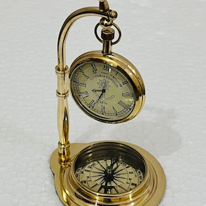 May include: A brass desk clock and compass set. The clock, marked "Victoria London 1876," is suspended above a compass. The compass is set in a circular base. The set is made of polished brass.