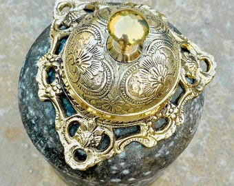 Puede incluir: Una campana de estilo antiguo con una parte superior dorada y detalles ornamentados. La campana se asienta sobre una base moteada gris y verde. La parte superior presenta un patrón floral y un pequeño mango redondo. El marco de la campana también es dorado.
